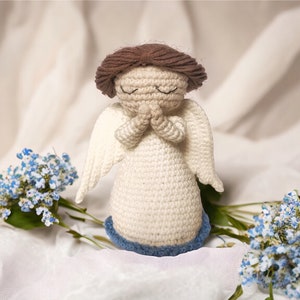 May include: A crocheted angel figurine with brown hair, white dress, and blue wings. The angel is holding its hands together in prayer. The figurine is sitting on a white fabric background with blue flowers.