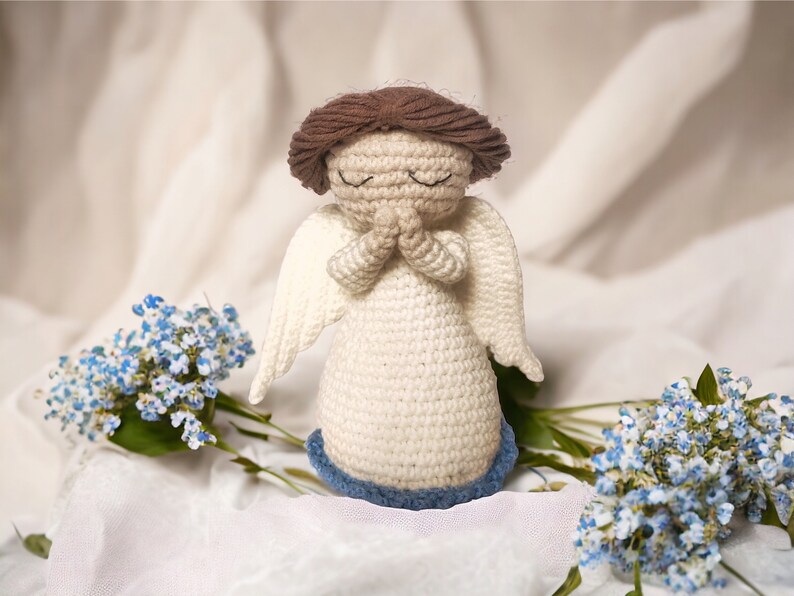 May include: A crocheted angel figurine with brown hair, white dress, and blue wings. The angel is holding its hands together in prayer. The figurine is sitting on a white fabric background with blue flowers.