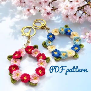 Könnte beinhalten: Zwei Schlüsselanhänger mit gehäkelten Blumenkränzen. Ein Kranz hat rosa und rote Blüten, der andere blaue und cremefarbene Blüten. Beide haben grüne Blätter und goldfarbene Schlüsselringe. Der Text "PDF pattern" ist in Blau geschrieben.