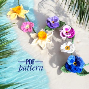 Op de afbeelding: Een gehaakte slinger met verschillende bloemen, waaronder gele narcissen, roze en paarse viooltjes en witte bloemen. De slinger ligt op een wit zandstrand met blauw water op de achtergrond. De tekst "PDF pattern" staat op de slinger.