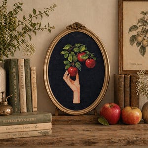 May include: An oval framed artwork with a gold frame features a hand reaching for red apples on a branch. The artwork is set on a wooden shelf with books, vases, and fresh apples. The overall theme is a still life.