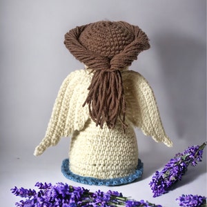 May include: A crocheted angel figurine with brown hair and white wings. The angel is standing on a blue and white base and is surrounded by lavender flowers.