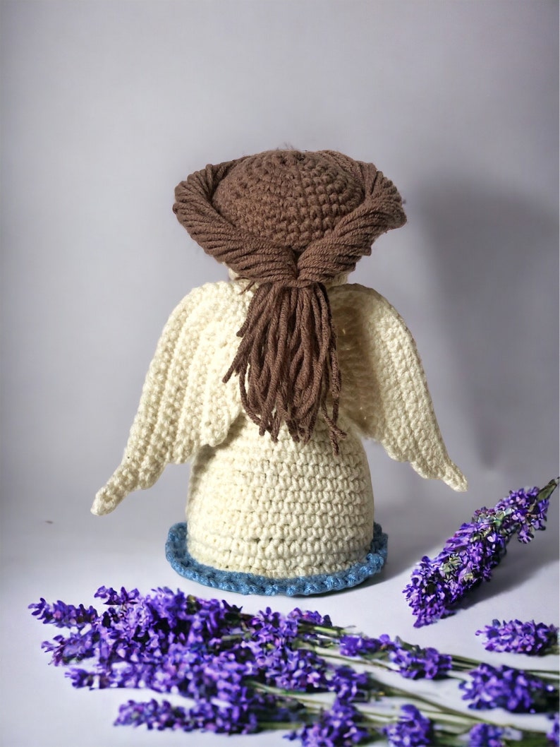 May include: A crocheted angel figurine with brown hair and white wings. The angel is standing on a blue and white base and is surrounded by lavender flowers.