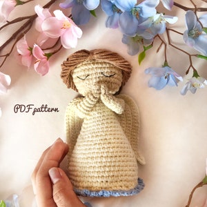 May include: A crocheted angel figurine with closed eyes and hands clasped in prayer. The angel is wearing a cream-coloured dress with a blue trim and has brown hair. The angel is surrounded by pink and blue flowers.