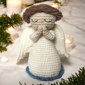 May include: A crocheted angel figurine made with white yarn and brown hair. The angel is wearing a white dress with blue trim and has its hands clasped in prayer.