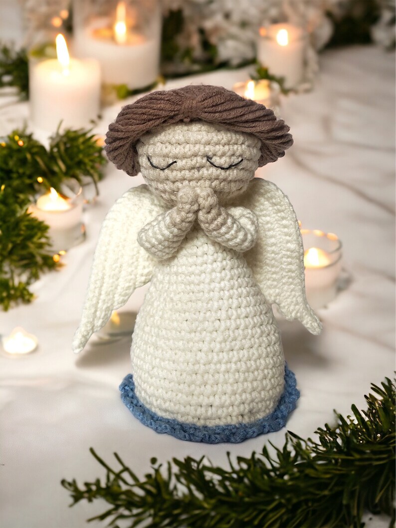 May include: A crocheted angel figurine made with white yarn and brown hair. The angel is wearing a white dress with blue trim and has its hands clasped in prayer.