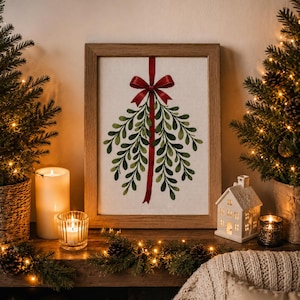 May include: A framed embroidery art piece featuring a green mistletoe sprig tied with a red ribbon and bow, centred on a light-coloured fabric background within a simple wooden frame, displayed as a decorative wall hanging for the holiday season.