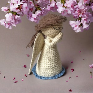 May include: A crocheted angel figurine with brown hair, white dress, and blue trim. The angel is standing with its hands clasped in prayer. Pink flower petals are scattered around the angel.