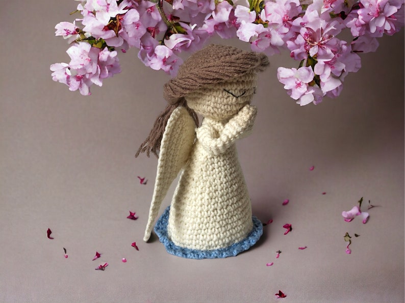 May include: A crocheted angel figurine with brown hair, white dress, and blue trim. The angel is standing with its hands clasped in prayer. Pink flower petals are scattered around the angel.