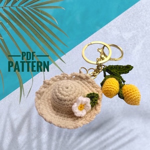 May include: A crochet keychain featuring a miniature beige sun hat with a white flower and green leaves. Attached is a gold-colored keyring and a crochet lemon branch with two yellow lemons. The text "PDF PATTERN" is visible.