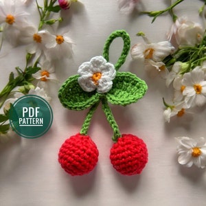 May include: A crocheted cherry charm with a green stem and leaves, a white flower with a yellow center, and two red cherries. The charm is on a white background with white flowers.