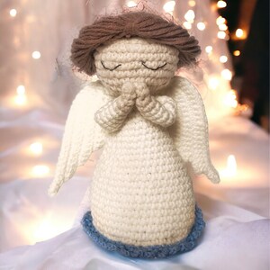 May include: A crocheted angel figurine made with white yarn and brown hair. The angel has its hands clasped in prayer and is wearing a blue trim around the bottom of its dress.