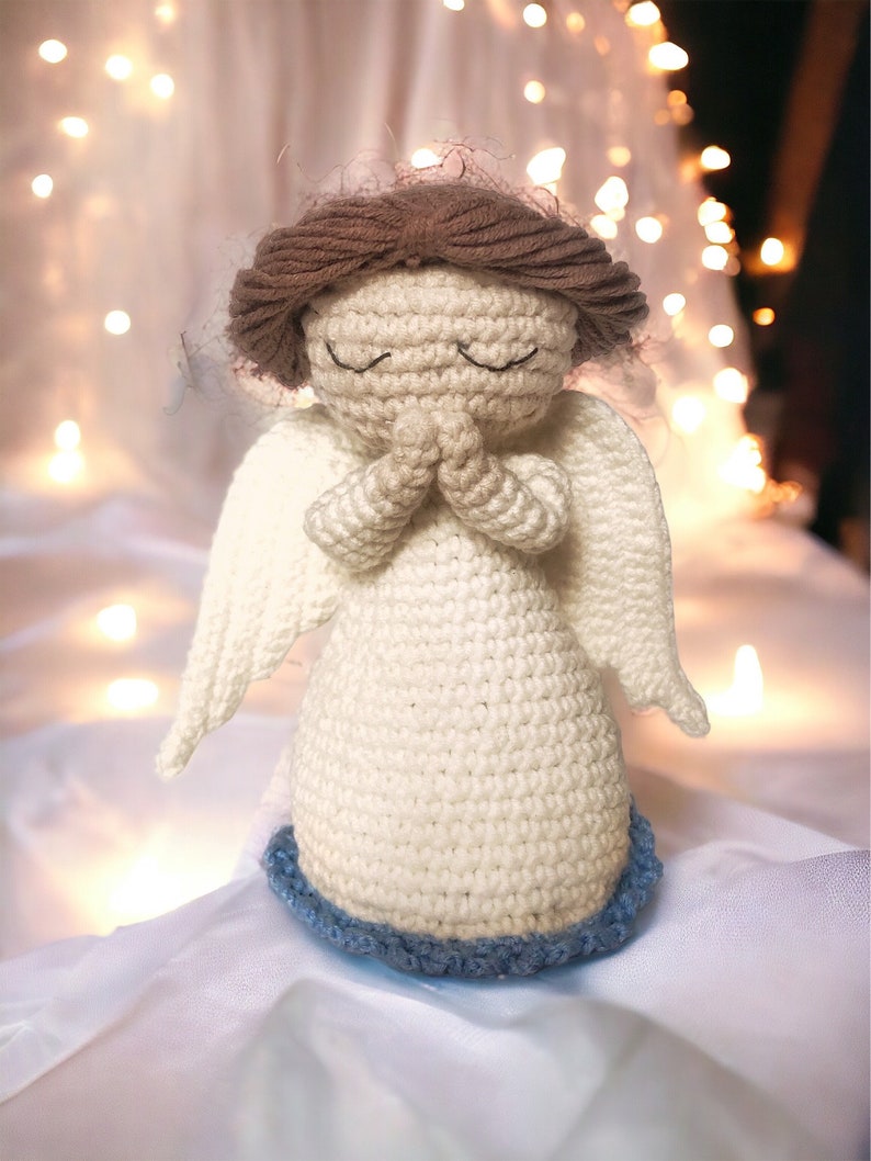 May include: A crocheted angel figurine made with white yarn and brown hair. The angel has its hands clasped in prayer and is wearing a blue trim around the bottom of its dress.