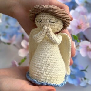 May include: A crocheted angel figurine with closed eyes and hands clasped in prayer. The angel is wearing a white dress with a blue trim and has brown hair.