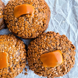May include: Three chocolate cookies topped with caramel and chocolate sprinkles. The cookies are on a white crumpled paper background.
