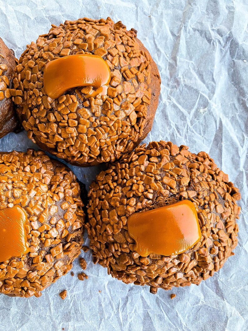 May include: Three chocolate cookies topped with caramel and chocolate sprinkles. The cookies are on a white crumpled paper background.