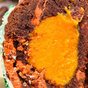 Pode incluir: Close-up de um muffin de chocolate cortado ao meio, revelando um recheio laranja brilhante. O exterior do muffin &eacute; marrom escuro com detalhes em laranja. Decorado com glac&ecirc; branco e granulado verde.
