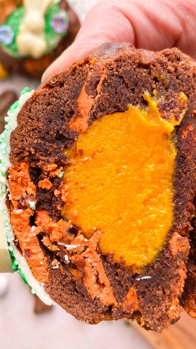 Pode incluir: Close-up de um muffin de chocolate cortado ao meio, revelando um recheio laranja brilhante. O exterior do muffin &eacute; marrom escuro com detalhes em laranja. Decorado com glac&ecirc; branco e granulado verde.