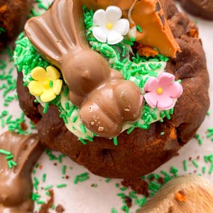 Pode incluir: Um donut de P&aacute;scoa de chocolate decorado com granulado verde, glac&ecirc; branco e decora&ccedil;&otilde;es de coelhinho e cenoura de chocolate. O donut &eacute; rodeado por outras guloseimas e granulado de chocolate.