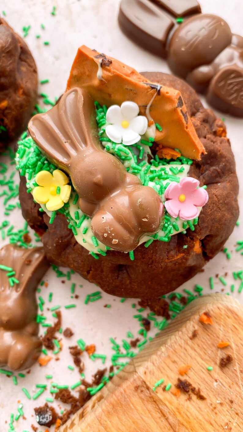 Pode incluir: Um donut de P&aacute;scoa de chocolate decorado com granulado verde, glac&ecirc; branco e decora&ccedil;&otilde;es de coelhinho e cenoura de chocolate. O donut &eacute; rodeado por outras guloseimas e granulado de chocolate.