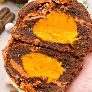 Pode incluir: Um bolo de P&aacute;scoa de chocolate cortado ao meio com um recheio laranja brilhante. O bolo tem um exterior castanho escuro com detalhes em laranja e branco. O topo &eacute; decorado com um coelhinho de chocolate branco e granulado verde. Ovos e coelhos de chocolate no fundo.