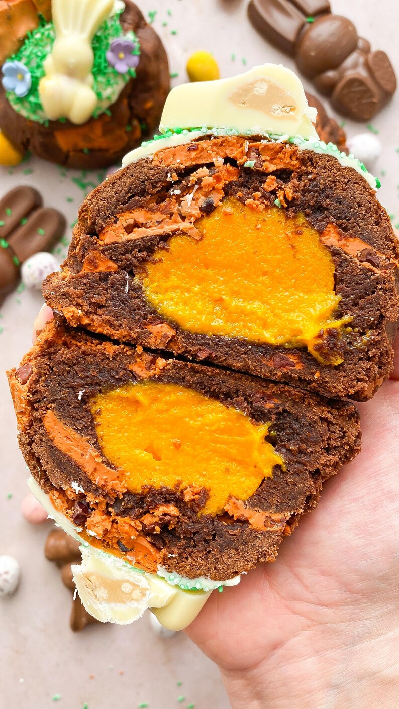 Pode incluir: Um bolo de P&aacute;scoa de chocolate cortado ao meio com um recheio laranja brilhante. O bolo tem um exterior castanho escuro com detalhes em laranja e branco. O topo &eacute; decorado com um coelhinho de chocolate branco e granulado verde. Ovos e coelhos de chocolate no fundo.