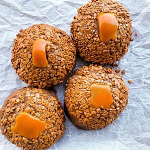 May include: Four chocolate cookies topped with caramel and chocolate sprinkles. The cookies are arranged on a white parchment paper background.