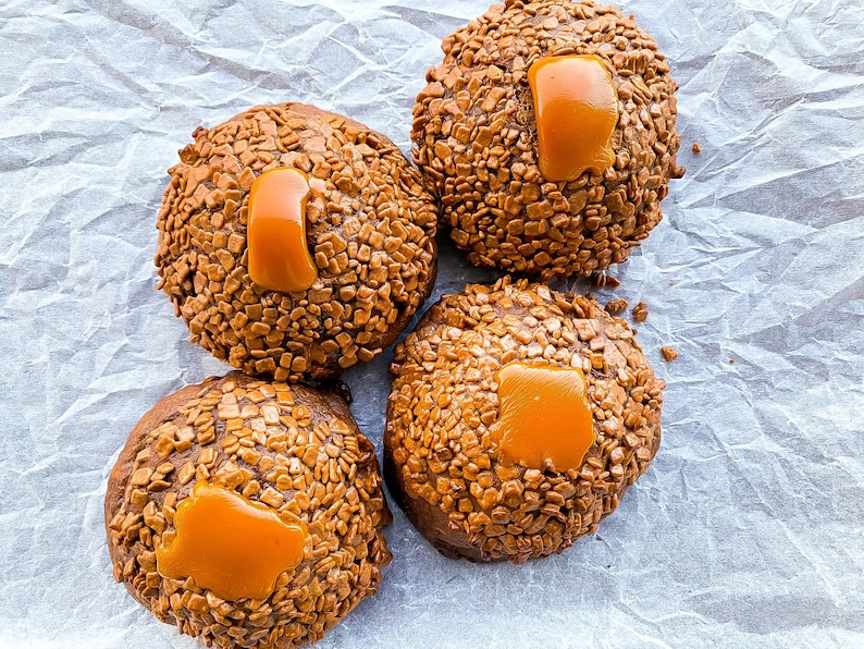 May include: Four chocolate cookies topped with caramel and chocolate sprinkles. The cookies are arranged on a white parchment paper background.