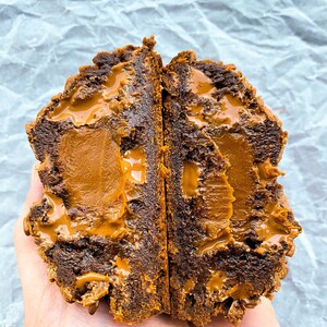 May include: A chocolate cookie filled with caramel and topped with a caramel drizzle. The cookie is broken in half, revealing the gooey caramel filling.
