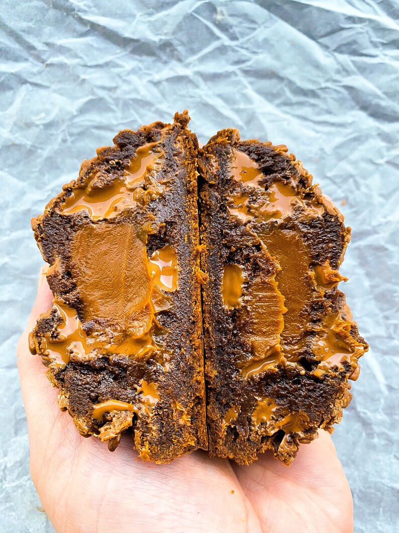 May include: A chocolate cookie filled with caramel and topped with a caramel drizzle. The cookie is broken in half, revealing the gooey caramel filling.