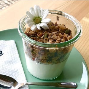 Può includere: Un barattolo di vetro riempito con strati di yogurt e granola, sormontato da una margherita bianca. Il barattolo è su un piatto verde, con un cucchiaio e un tovagliolo in primo piano. La granola ha un mix di semi e noci.