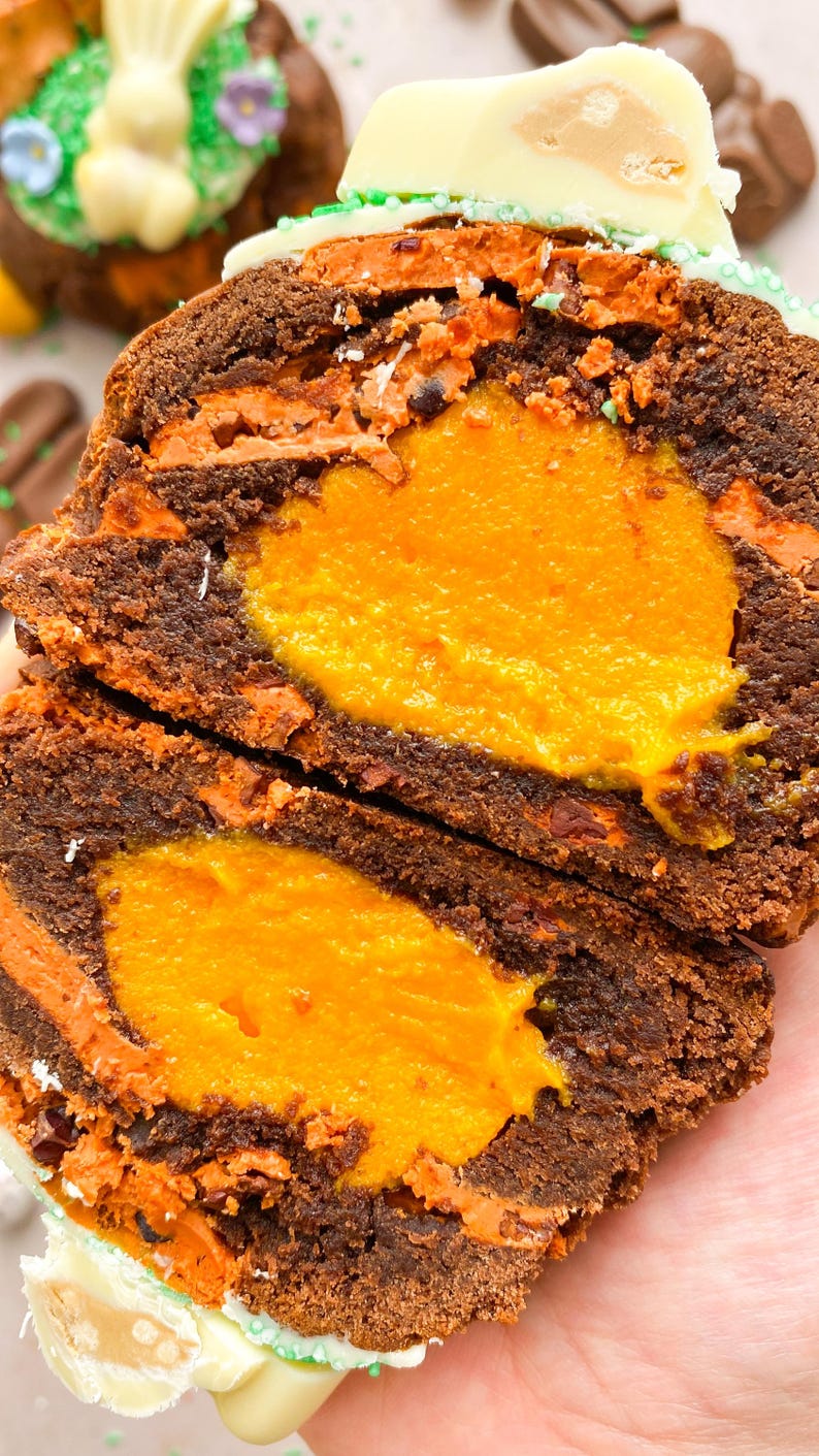 Pode incluir: Close-up de uma sobremesa de chocolate cortada ao meio com um recheio laranja brilhante. A sobremesa tem camadas de bolo marrom escuro e cobertura laranja. Uma pe&ccedil;a de chocolate branco com um centro marrom claro fica em cima.