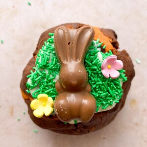 Pode incluir: Um coelho de P&aacute;scoa de chocolate em cima de um pastel de chocolate, decorado com granulado verde e doces em forma de flor rosa e amarela. O pastel tem uma base de glac&ecirc; branco.