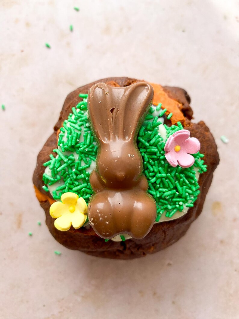 Pode incluir: Um coelho de P&aacute;scoa de chocolate em cima de um pastel de chocolate, decorado com granulado verde e doces em forma de flor rosa e amarela. O pastel tem uma base de glac&ecirc; branco.