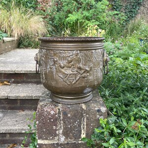 May include: A large, ornate brass planter with intricate carvings and two ring handles. The planter is sitting on a brick wall.