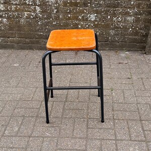 May include: A square stool featuring a bright orange seat with a mottled texture and a black metal frame. The frame consists of four tubular legs connected by horizontal support bars on all sides, providing a stable base for the seat.