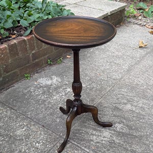 May include: A dark brown wooden pedestal table featuring a circular top with a visible wood grain pattern, a slender central column with carved detailing, and three curved legs that extend outward to provide a stable base for the furniture piece.