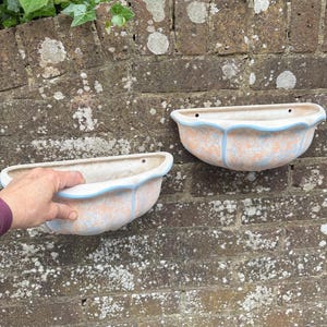 May include: Two wall-mounted planters with a scalloped edge and a light blue trim. The planters have a peach and white mottled finish. They are mounted on a weathered brick wall.
