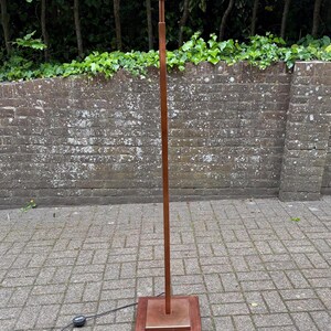 May include: A tall, slender floor lamp with a copper finish. The lamp has a square base, a long, straight pole, and a small, cylindrical top. The lamp is approximately 152 cm tall and stands on a brick patio.