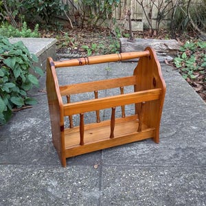 May include: A light brown wooden magazine rack. The rack has a rectangular base, vertical supports, and a handle at the top. The design features a series of vertical spindles and horizontal slats for holding magazines or books. The wood grain is visible.