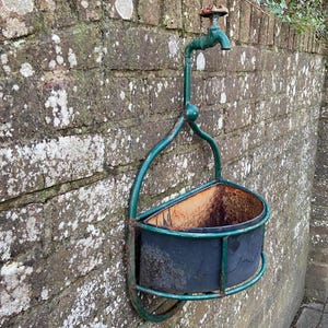 May include: A green metal planter with a rusty metal pot inside. The planter is attached to a brick wall with a green metal tap.