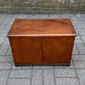 May include: A rectangular wooden chest with a rich, warm brown finish. The chest features a flat top and two doors with a chevron pattern. The chest is approximately 76 cm wide, 46 cm deep, and 61 cm tall.
