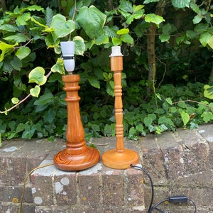 May include: Two wooden table lamps with white lampshades. The lamps are on a brick wall with green foliage in the background.