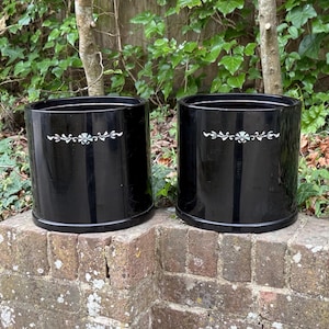 May include: Two black, cylindrical plant pots with a glossy finish. Each pot has a white, floral design near the top. The pots are on a weathered brick surface, with greenery in the background.