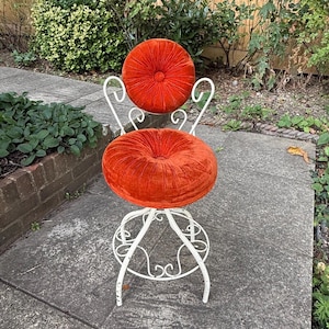May include: A swivel stool featuring a white metal frame with scrollwork details and two round, tufted cushions upholstered in a vibrant orange velvet fabric. The seat and backrest are circular, providing a distinct contrast to the white base.