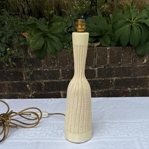 May include: A cream-coloured table lamp with a textured, carved design. The lamp has a slender, elongated shape with a brass-coloured bulb fixture. The lamp's cord is visible, and the background includes greenery and a brick wall.