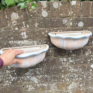 May include: Two ceramic wall planters with a scalloped edge and a light blue trim. The planters have a speckled peach and white finish. The planters are mounted on a weathered brick wall.
