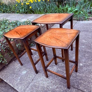 May include: Three wooden side tables with square tops and angled legs. The tables have a rich, warm brown finish, showcasing the natural wood grain. The tables are arranged outdoors, suggesting a patio or garden setting.