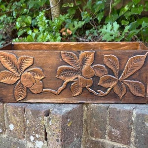 May include: A rectangular, brown planter box with embossed chestnut leaf and nut designs. The planter is made of a dark brown material and sits on a brick wall, with green foliage in the background. The box is approximately 30 cm long.