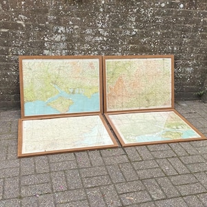 May include: Four rectangular framed maps featuring detailed topographical cartography with green, tan, and blue colour palettes. Each map is encased in a simple, thin wooden frame, displaying various land features, coastlines, and grid lines.